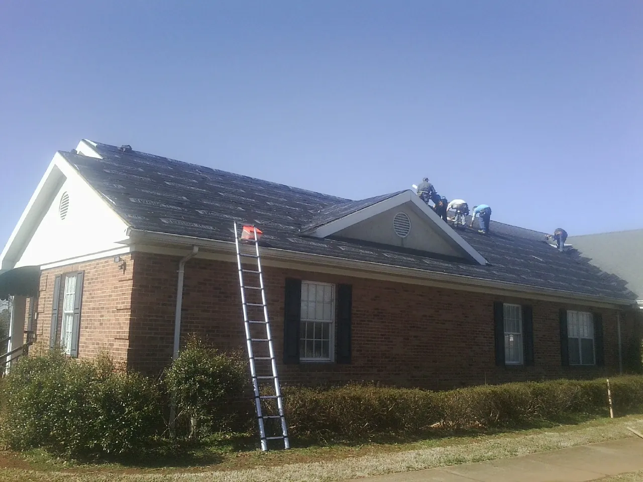 Crew tearing shingles off a brick ranch roof with a ladder set — Emergency Roof Repair work by our crew in Floral Park