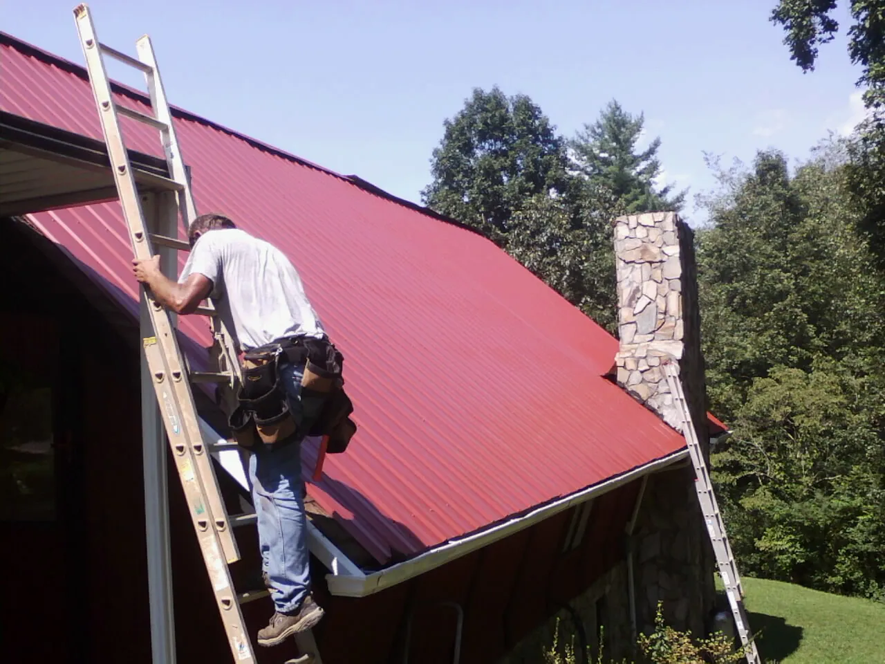 Roofer on a ladder beside a red metal roof and stone chimney — Soffit & Fascia Repair by Roofing Brothers in Floral Park